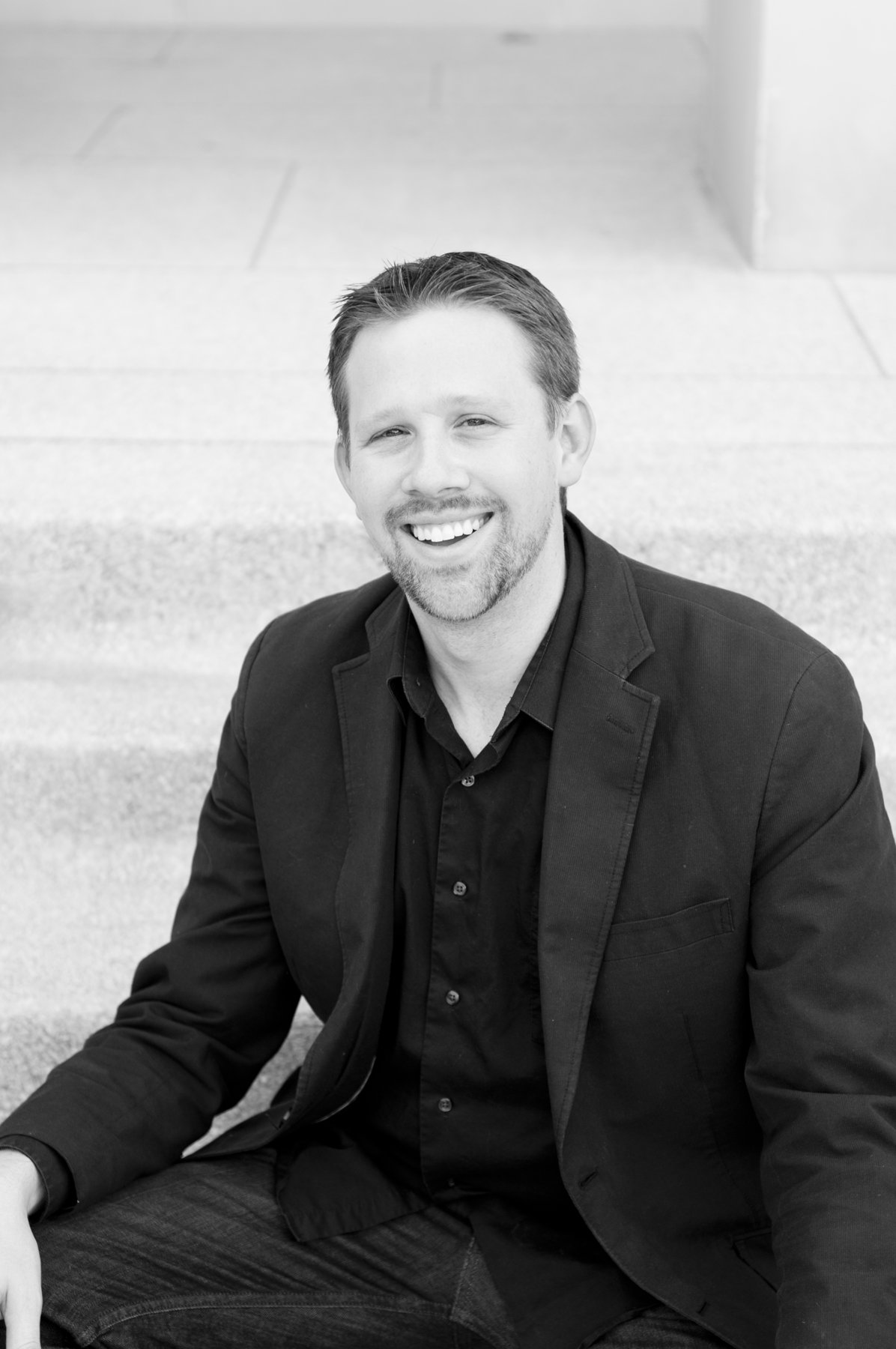 Ryan Ross, smiling, seated on stone steps in a dark blazer